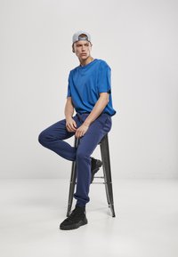 Modrá bavlnená tričko, námornícke tepláky a čierne športové topánky; usadený na kovovom stoličke, nosiaci šedú čiapku. Čistý dizajn, minimálne detaily.