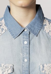 Chemise en denim bleu clair avec un col pointu, ornée de détails en dentelle crème sur les épaules et de deux poches à bouton sur la poitrine.