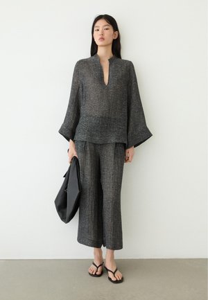 Vrouw die losse, donkergrijze, gestructureerde wijde broek draagt en een bijpassende top met diepe V-hals, een zwarte tas vasthoudend, staand tegen een witte muur.