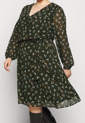 Vrouw draagt een donkergroene knielange jurk met meerkleurige bloemenprint, lange doorschijnende mouwen, een gestrikte taille en zwarte kniehoge laarzen.