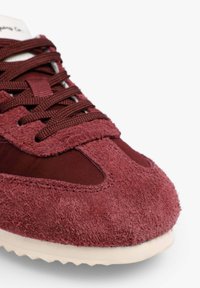 Zapatilla de ante burdeos con cordones a juego, detalles lisos y puntera texturizada. Presenta una suela ligera con un patrón de dibujo.