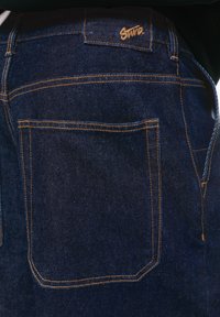 Donkerblauwe spijkerbroek met een standaard achterzak met contrasterend oranje garen en een klein geborduurd logo boven de zak.