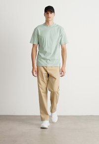 Homem a caminhar, a usar um chapéu preto, uma t-shirt verde-claro, calças bege e ténis brancos, contra um fundo branco liso.
