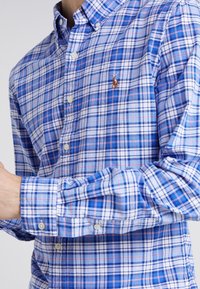 Koszula w kratkę w kolorze niebieskim, białym i czerwonym, wykonana z tkaniny tkanej. Posiada kołnierzyk zapinany na guziki oraz haftowane logo po lewej stronie klatki piersiowej.