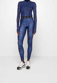 Koko kehon urheiluasu, jossa on violetti ja sininen kuvioitu pitkähiha toppi ja siihen sopivat leggingsit, mustalla brändätyllä vyötärönauhalla, yhdistettynä valkoisiin lenkkareihin.