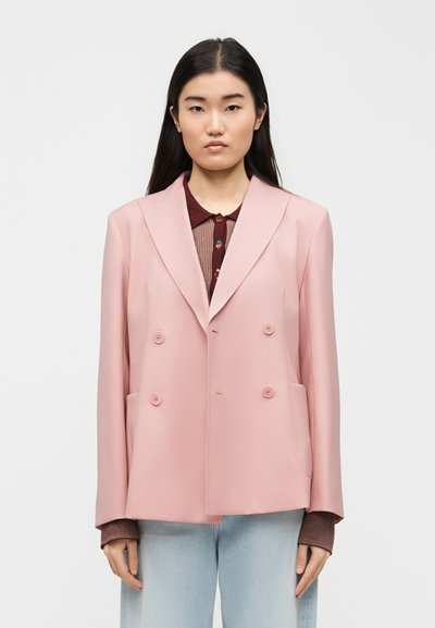 Young woman with long dark hair wearing a light pink double-breasted blazer over a brown top and light blue jeans, standing against a white background.