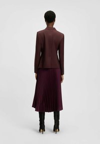 Bordowy żakiet dopasowany w połączeniu z plisowaną spódnicą. Żakiet ma gładkie linie, a spódnica prezentuje pionowe plisy, obie z delikatnym połyskiem.