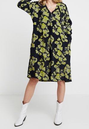 Frau trägt ein loses schwarzes Kleid mit gelbem Blumenmuster und weißen Stiefeletten, steht mit einer Hand hinter dem Kopf und der anderen in der Tasche.