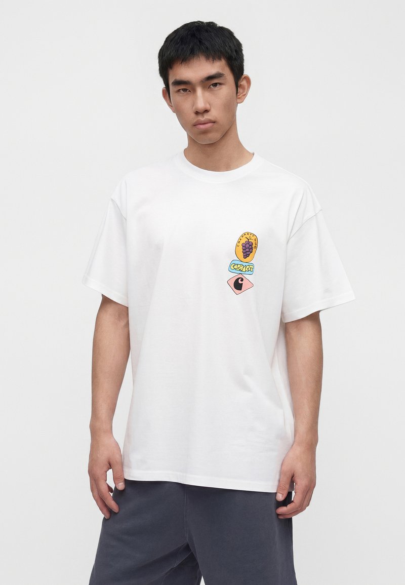 Junger Mann trägt ein weißes übergroßes T-Shirt mit kleinen bunten Trauben- und Textgrafiken auf der linken Brust, kombiniert mit dunkler Hose.