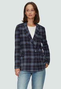 Marine geblokte blazer met blauw-witte ruitpatroon, dubbelgeknoopte ontwerp, met notch revers en twee voorkruiszakken. Draagt over een witte top.