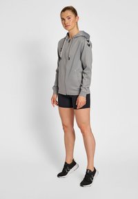 Felpa grigia con zip e cappuccio, con accenti a strisce nere sulle spalle. Abbinata a pantaloni corti neri e scarpe da ginnastica nere.