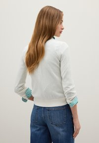 Witte geribde trui met groene en witte gestreepte manchetten. Aansluitend ontwerp, middelzware textuur en geribde zoom. Geko, met blauwe jeans.