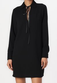 Robe noire avec un col, des manches longues et un décolleté en forme de clé. Présente un lacet fin et une texture de tissu lisse.