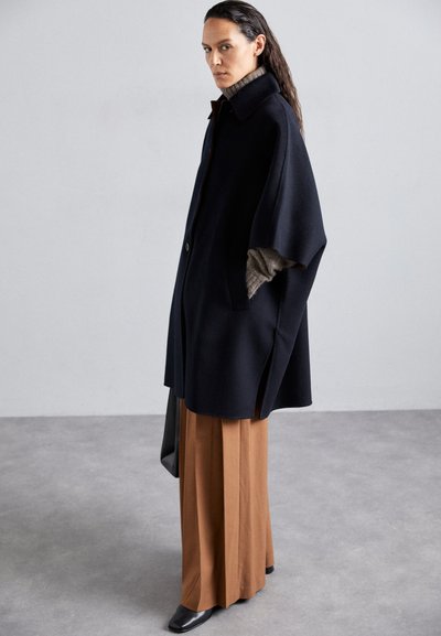 Cappotto oversized blu navy con colletto alto, chiusura con bottoni e spacchi laterali. Indossato sopra pantaloni marroni a gamba larga e abbinato a scarpe nere a punta.