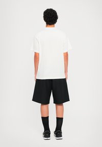 Άτομο με σγουρά μαλλιά, φορώντας ένα απλό λευκό μπλουζάκι, μαύρες σορτς μέχρι το γόνατο, μαύρες κάλτσες και sneakers σε μαύρο και άσπρο, που στέκεται στραμμένο προς τα πίσω.