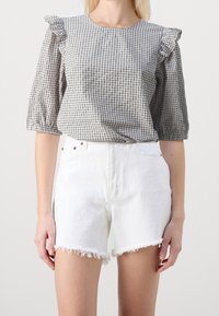 Donna che indossa una blusa a scacchi bianco e nero con spalle arricciate e jeans shorts bianchi a vita alta con orli sfilacciati.