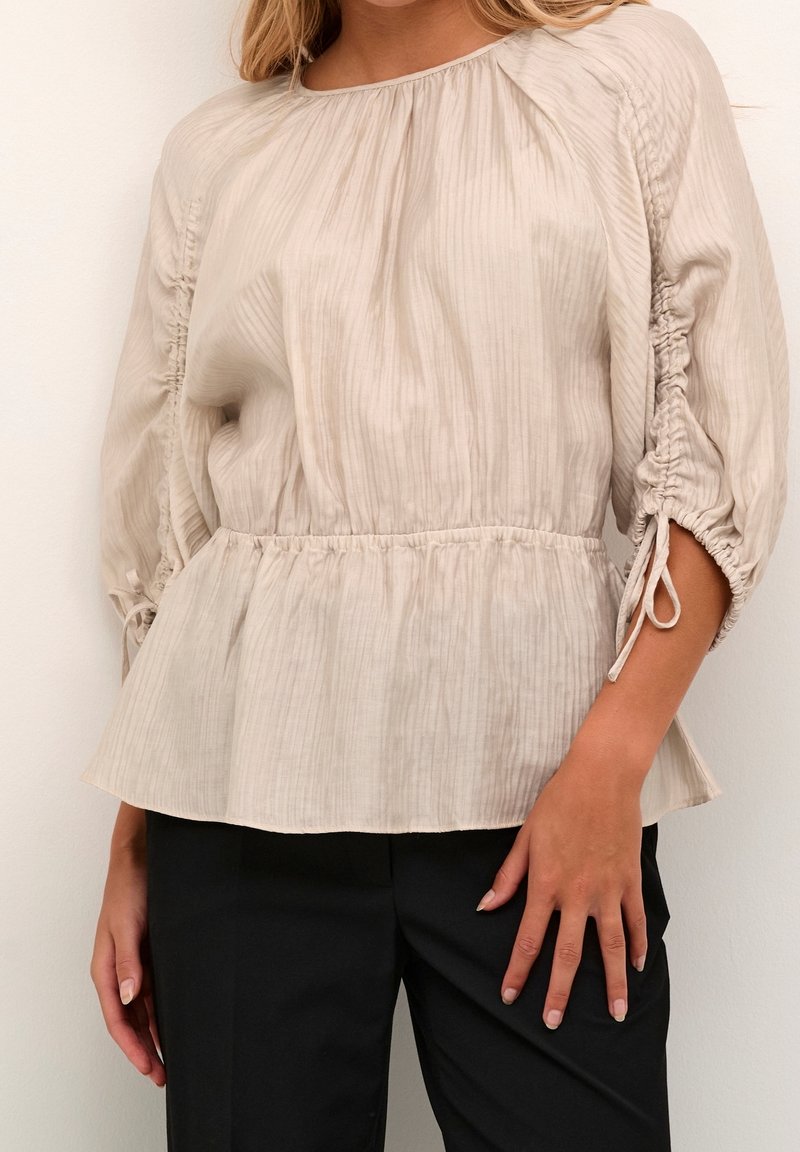 Femme portant un chemisier beige texturé avec des manches 3/4 froncées et une taille peplum, assorti à un pantalon noir sur un fond uni.
