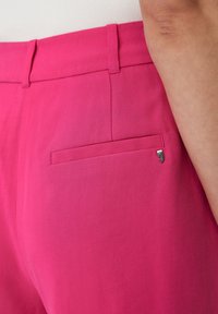 Primo piano di una persona che indossa dei pantaloni rosa acceso con passanti per cintura e una tasca posteriore arricchita da una piccola spilla a forma di virgola argentata.