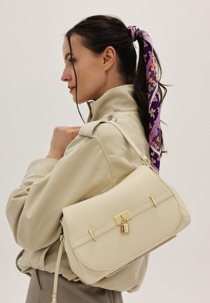 Frau mit dunklem Haar, das in einem lila floralen Schal gebunden ist, trägt eine beige Jacke und hat eine cremefarbene Handtasche mit einem goldenen Verschluss auf der Schulter.