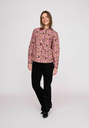 Ung kvinde smiler, står op, iført en lyserød blomstret langærmet skjorte, sorte bukser og sorte spidse sko mod en ensfarvet lys baggrund.