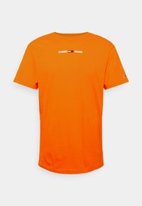 Pomarańczowa bawełniana koszulka z krótkim rękawem, okrągłym dekoltem. Zawiera biały napis "TOMMY JEANS" na wysokości klatki piersiowej. Prosty, jednolity design.