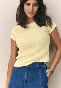 Haut jaune texturé à manches courtes avec un col rond, associé à un jean taille haute bleu. Présente de légères rides dans le tissu.