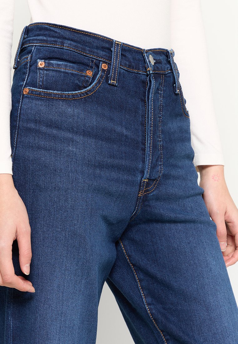 Jeans in denim blu a vita alta, con rivetti in rame, cinque tasche e diverse cuciture arancioni. Il tessuto appare liscio e resistente.