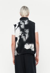 Melna un balta polārzvīņa veste ar abstraktu rakstu, augstu apkakli un bez piedurknēm. Modelis to valkā pār baltu krekli.
