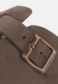 Detail eines braunen Wildledersandalen mit einem breiten Riemen und einer Metallnadel. Die Oberfläche ist weich und hat ein leicht strukturiertes Finish.