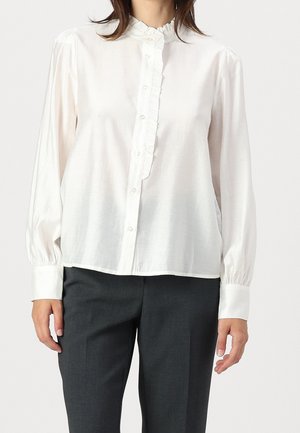 Femme portant un chemisier blanc à manches longues avec des boutons et des volants, associé à un pantalon gris foncé.