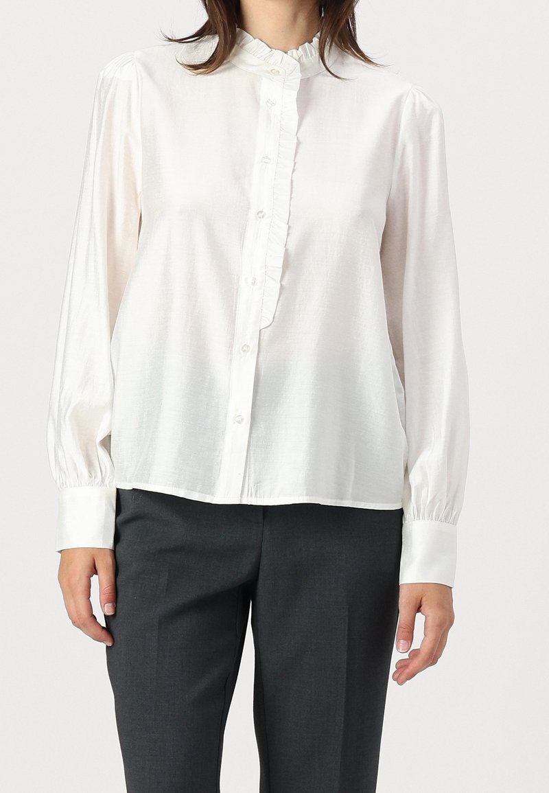 Femme portant un chemisier blanc à manches longues avec des boutons et des volants, associé à un pantalon gris foncé.