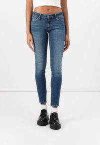 Közép-kék farmer skinny nadrág, normál derékrésszel, elülső zsebekkel és enyhe kopással. Fekete magassarkú cipővel párosítva.