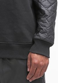 Mano destra di una persona che indossa una giacca di pelle trapuntata nera con polsini a coste e pantaloni neri, su uno sfondo bianco.