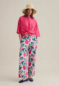 Chemise rose à manches longues nouée à la taille, associée à un pantalon large à fleurs dans les tons rose, vert et bleu, complétée par des ballerines bleu marine et un chapeau en paille.