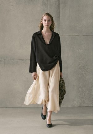 Frau geht drinnen entlang, trägt eine lockere schwarze Bluse, einen fließenden beigen Rock, schwarze Ballerinas und hält eine gemusterte Tasche vor einer grauen Wand.