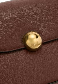 Pochette in pelle marrone con texture e una chiusura circolare in metallo dorato molto visibile, bordatura nera. Presenta una finitura liscia e un marchio discreto.