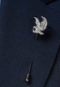 Hopeaket paluu muotoinen hopeapinni, joka on yksityiskohtaisesti teksturoitu, kiinnitetty tummansiniselle kankaalle. Pieni musta neula näkyy kiinnittämistä varten.