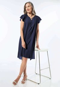 Navy-blaues A-Linien-Kleid mit kurzen Rüschärmeln, Falten und V-Ausschnitt. Trägt dazu helle, hohe Sandalen vor einem neutralen Hintergrund.