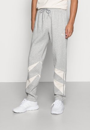 Personne portant un pantalon de survêtement gris clair avec des panneaux géométriques blancs et des baskets blanches, debout devant un fond clair uni.