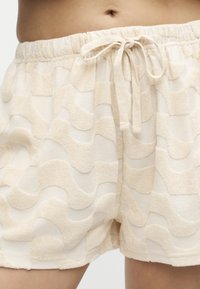 Μέση και άνω πόδια ατόμου που φοράει μπεζ σορτς από υφασμάτινο ύφασμα με κυματιστό μοτίβο και ρυθμιζόμενο κορδόνι στη μέση.