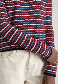 Gebreide pullover met een rood, blauw en wit horizontaal patroon. Beschikt over een golvende zoom en manchetten, gemaakt van een zachte, gestructureerde stof.