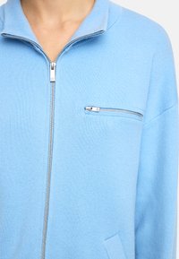 Jasnoniebieski sweter na zamek z żebrowanym wzorem, stójką oraz kieszenią na zamek na klatce piersiowej. Posiada luźny krój oraz kieszenie po bokach.