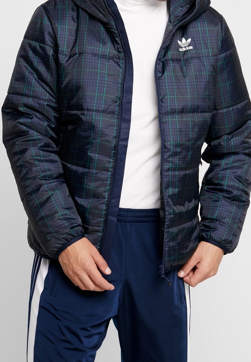Navy puffer jacket with a plaid pattern in green and black. Features a zip closure, hood, and two side pockets. Reflective Adidas logo.