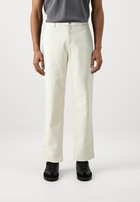 Pantaloni albi din bumbac cu un design drept, dotati cu închidere cu nasture în față și bucle pentru curea, purtați cu adidași negri.