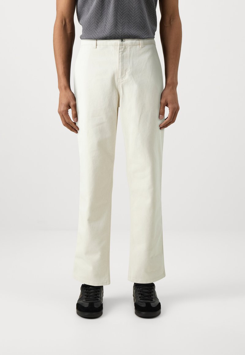 Pantalones de algodón blancos con un diseño de pierna recta, que cuentan con un cierre frontal de botón y presillas para cinturón, combinados con zapatillas negras.
