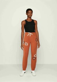 Calças de treino laranja com padrões florais em branco, castanho e rosa. Apresenta um cós elástico com cordão. Combinadas com uma t-shirt preta.