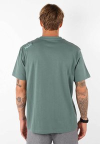 T-shirt en coton vert avec un col rond, des manches courtes et un patch logo dans le haut du dos. Texture lisse avec une coupe décontractée.