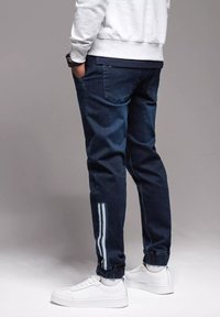 Pantalones jogger azul oscuro con puños elásticos, con rayas laterales blancas y un diseño casual. Combinados con zapatillas blancas.
