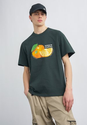 Mladý muž v čiernej čiapke, tmavozelenom tričku Nike s oranžovou a citrónovou grafikou a béžových cargo nohaviciach, stojaci pred jednofarebným pozadím.