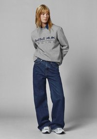 Sudadera gris con el logo de "Red Bull Racing", jeans de mezclilla azul y zapatos deportivos blancos. Cabello peinado lacio, suelto. Fondo sencillo.
